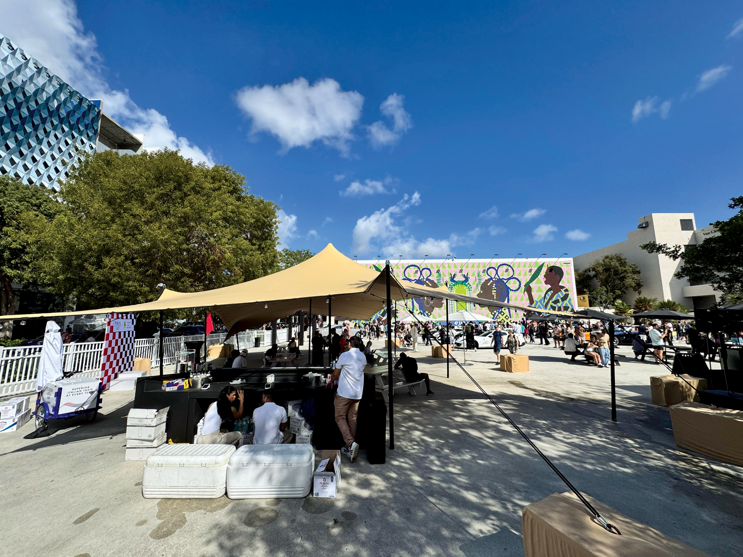 A vibrant outdoor gathering with a large mural, tents for vendors, and a crowd enjoying the day under a bright blue sky.