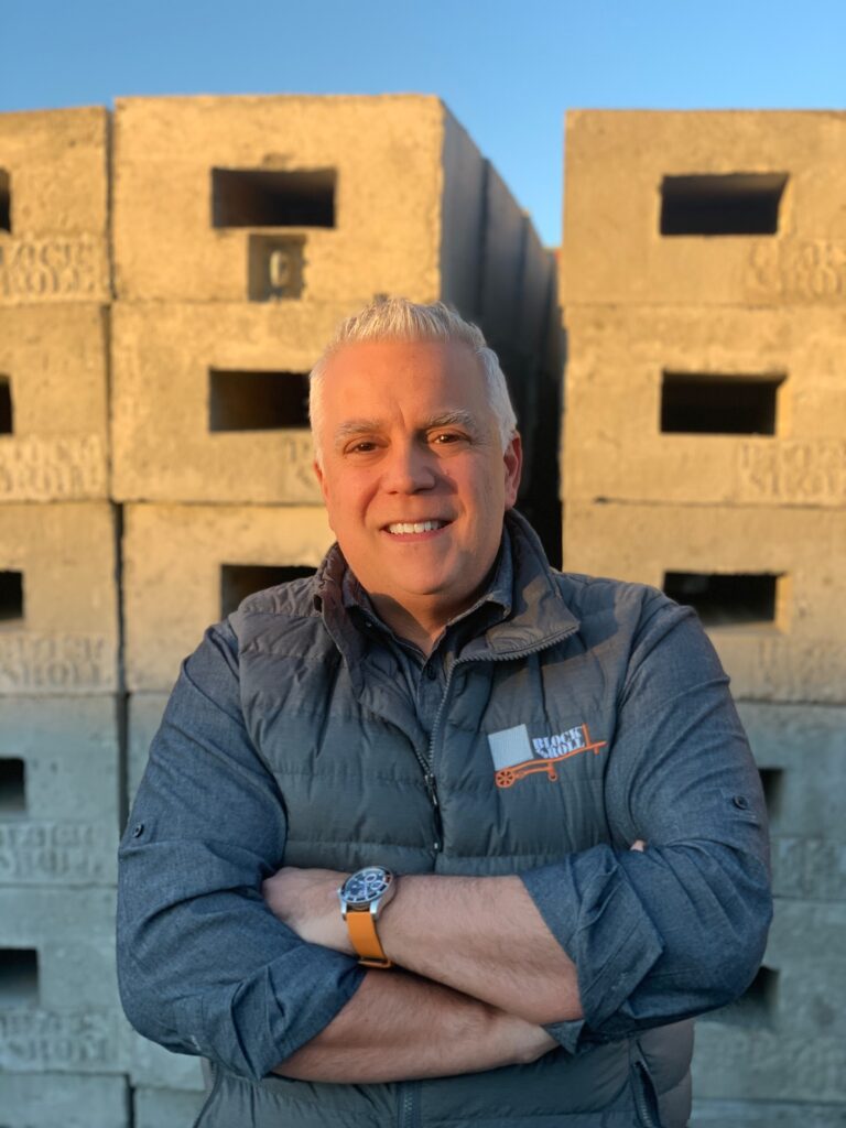 NIck A man with short, light hair smiles confidently, standing with arms crossed in front of stacked concrete blocks, under a clear sky.