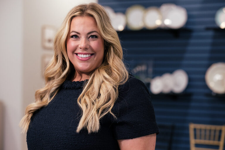 HTE A smiling woman with long, wavy blonde hair, wearing a dark top, stands against a blue wall with white plates displayed on shelves.