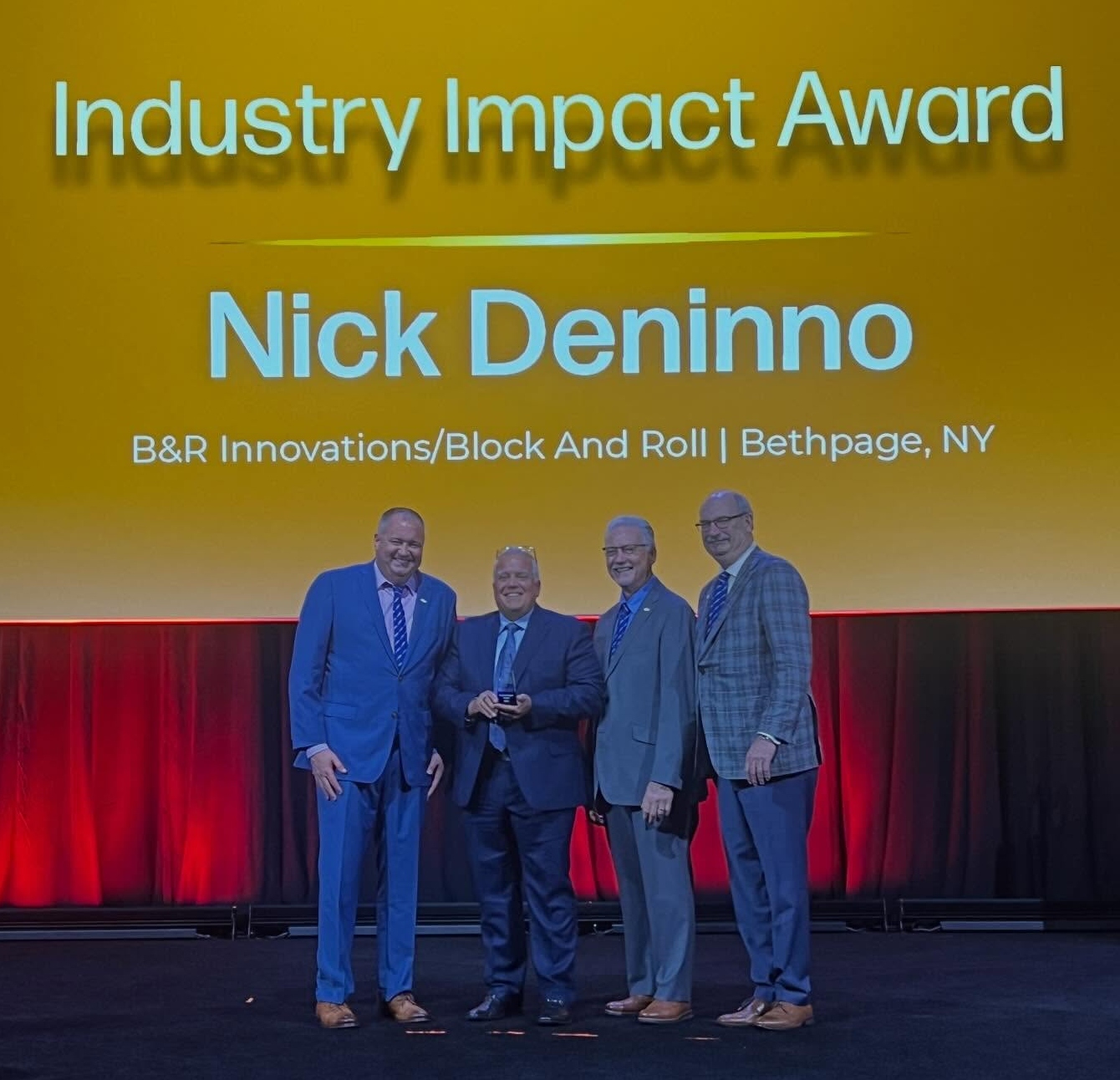 Deninno ARA Award RESIZED Four men in formal attire stand on stage under a bright yellow backdrop, celebrating Nick Deninno with an award.